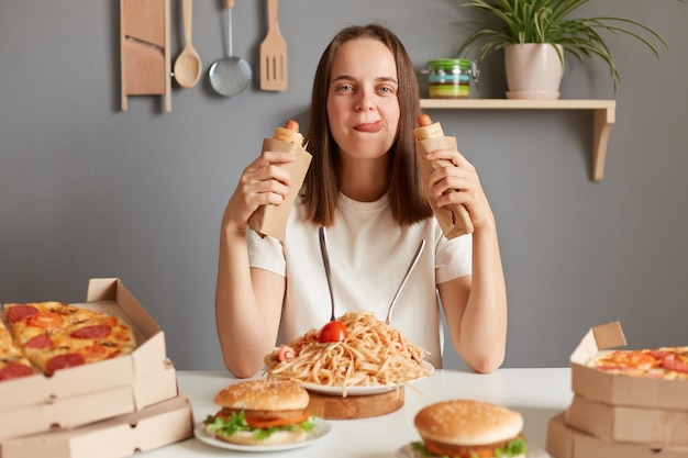 Retrato de una mujer hambrienta con el pelo castaño y una camiseta blanca sentada en la mesa comiendo salchichas en masa mostrando la lengua fuera con un trastorno alimentario comiendo comida chatarra después de la dieta