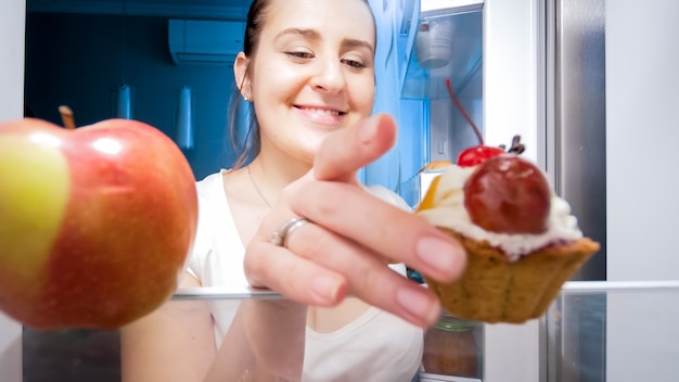 Retrato de mujer hambrienta tomando pastel dulce en lugar de manzana helthy para bocadillos por la noche. Concepto de dieta y nutrición saludable.