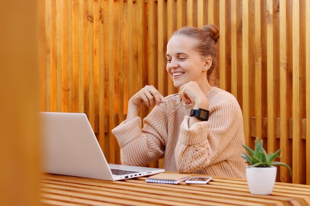 Retrato de una mujer hermosa, agradable y sonriente, con peinado de moño y suéter beige, trabajando en una laptop contra una pared de madera, mirando la pantalla del portátil y sosteniendo anteojos ópticos en las manos