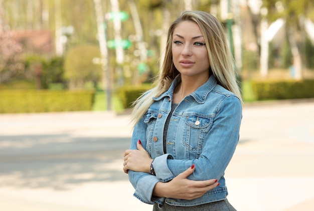 Elegante mujer joven hermosa con maquillaje natural con labios sexy en una  chaqueta de mezclilla azul con un vestido negro con un bolso de moda se  encuentra en la ciudad en un