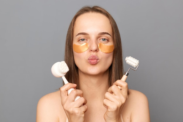 Retrato de mujer hermosa alegre romántica con cabello oscuro sosteniendo rodillo de masaje facial y cepillo de pie con parches debajo de los ojos enviando besos de aire posando contra la pared gris