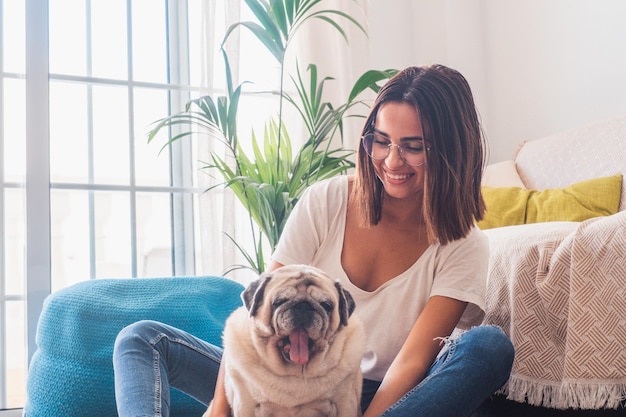 Retrato de una mujer hermosa con anteojos divirtiéndose con su perro pug sentado en el suelo en la sala de estar de su casa Mujer alegre pasando tiempo libre con su lindo perro mascota en casaxA