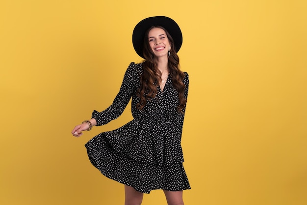 Retrato de una mujer hermosa y atractiva posando aislada en un fondo amarillo con un vestido negro punteado y un sombrero negro con estilo accesorios de estilo de moda de primavera verano sonriendo feliz humor divirtiéndose