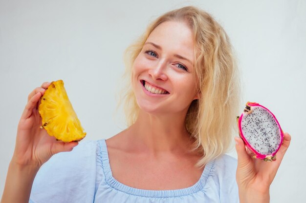 Retrato de mujer hermosa de cabello rubio con ojos azules sosteniendo piña y fruta de dragón sobre fondo blanco desintoxicación y alergia a productos tropicales