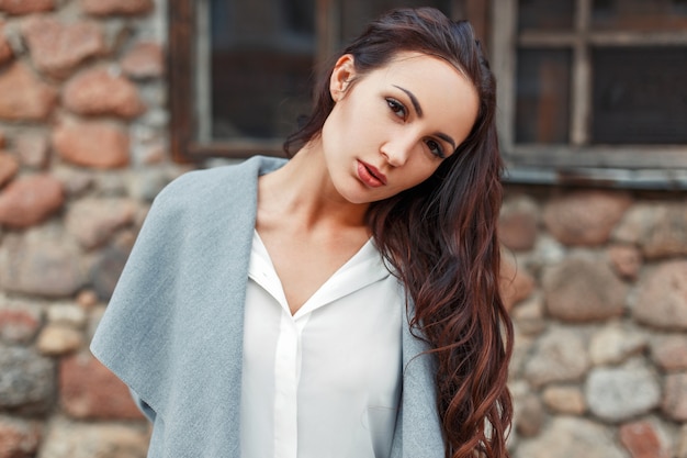 Retrato de una mujer hermosa con estilo en un abrigo gris y una blusa blanca contra un muro de piedra