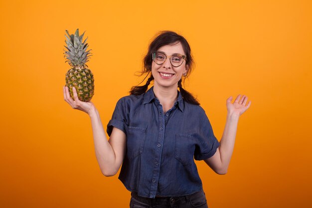 Retrato de una mujer hermosa con gafas sonriendo a la cámara y sosteniendo unas deliciosas ananas en el estudio sobre un fondo amarillo. Mujer caucásica feliz con frutas exóticas de verano.