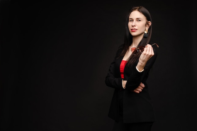 Retrato de mujer hermosa joven mirando pensativamente hacia abajo Modelo morena en traje elegante posando en la cámara Señora bonita en gafas de sol rojas apoyando una mano sobre la otra y sosteniéndola cerca de la cara