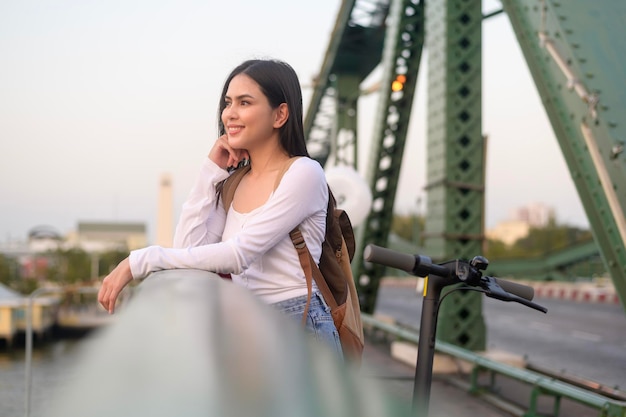 Retrato de mujer hermosa joven con un scooter eléctrico sobre el puente en el fondo de la ciudad moderna