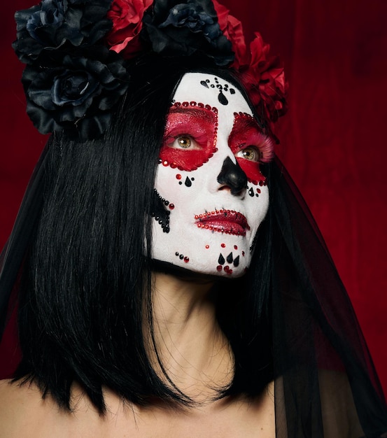 Retrato de una mujer hermosa con un maquillaje de calavera de azúcar con una corona de flores en la cabeza, fondo rojo.