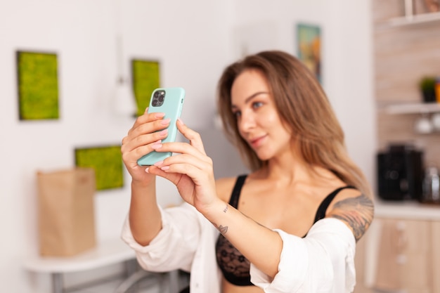 Retrato de mujer hermosa mirando a la cámara del teléfono mientras toma selfie en la cocina de casa con ropa interior sexy. Mujer seductora con tatuajes con smartphone usando lencería temping en la mañana.