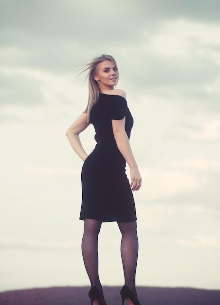 Retrato de una mujer hermosa modelo de moda posando en un elegante vestido negro sobre fondo de cielo azul Tiempo nublado soleado Disparo al aire libre