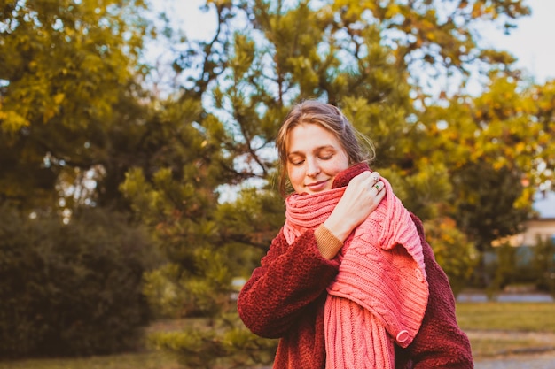 Retrato de una mujer hermosa con los ojos cerrados envolviéndose en una cálida bufanda de punto rosa. Chica soñadora en bufanda rosa brillante hecha a mano con parque de otoño en el fondo.