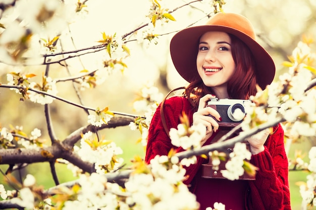 Retrato de una mujer hermosa pelirroja en suéter rojo y sombrero con cámara en jardín de manzano en flor en primavera al atardecer.