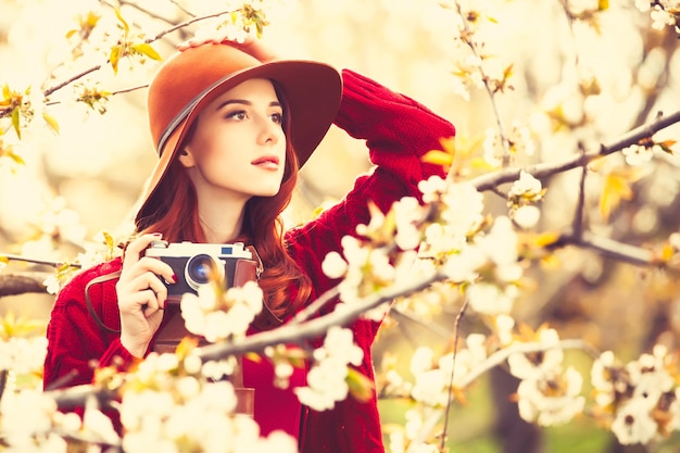 Retrato de una mujer hermosa pelirroja en suéter rojo y sombrero con cámara en jardín de manzano en flor en primavera al atardecer.