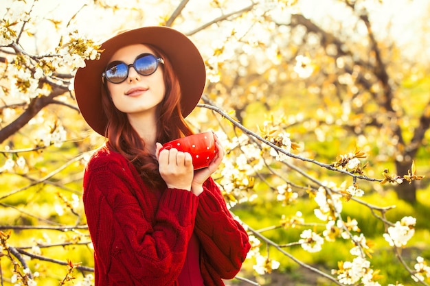 Retrato de una mujer hermosa pelirroja en suéter rojo y sombrero con taza en jardín de manzano en flor en primavera al atardecer.