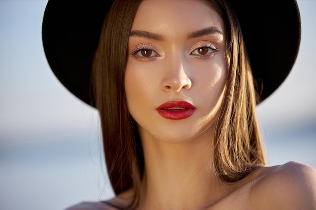 Retrato de una mujer hermosa con el pelo largo en un gran sombrero negro redondo. Rostro de piel de belleza, hombros desnudos. Chica luz dorada al atardecer