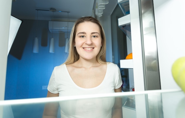 Retrato de mujer hermosa en pijama mirando dentro del refrigerador por la noche