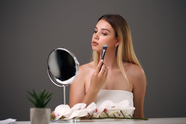 Retrato de una mujer hermosa con pincel de maquillaje cerca de su rostro y sonriendo en gris