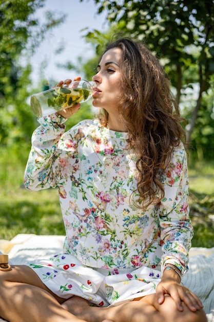 Retrato de mujer hermosa posando acostada sobre tela escocesa en el parque del jardín de verano un libro de frutas una bolsa y un agua...