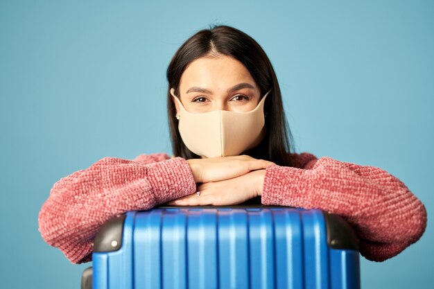 Retrato de mujer hermosa posando con maleta y máscara facial, aislado sobre fondo azul. Copie el espacio. Concepto de viaje, coronavirus.