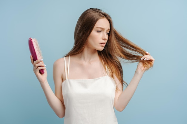 Retrato de una mujer hermosa y preocupada en camiseta sosteniendo un cepillo de pelo aislado sobre un fondo azul