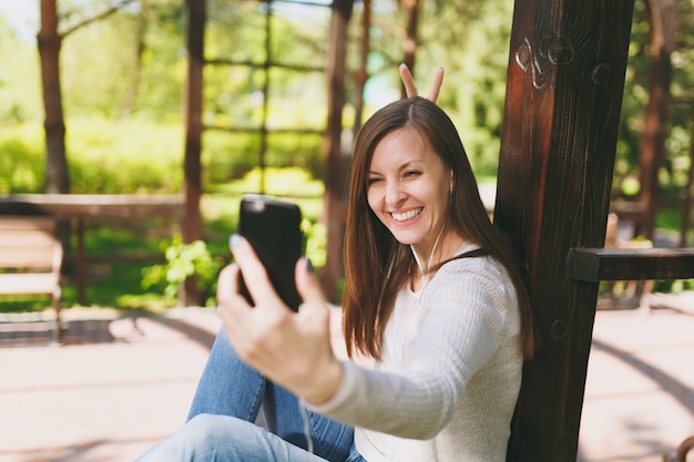 Retrato de mujer hermosa con ropa casual ligera. Mujer sonriente sentada en el parque de la ciudad en la calle al aire libre en la naturaleza de primavera, haciendo selfie en teléfono móvil o videollamada. Concepto de estilo de vida.