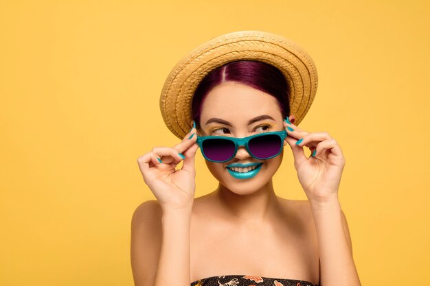 Retrato de mujer hermosa con sombrero de maquillaje brillante y gafas de sol sobre fondo amarillo studio