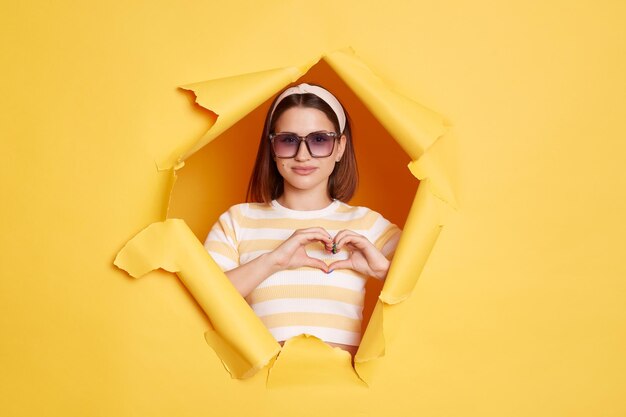 Retrato de una mujer hermosa sonriente con pantalones a rayas y una banda para el pelo posando en un agujero de papel amarillo mirando a la cámara mostrando el símbolo de amor hace un gesto de forma de corazón con las manos