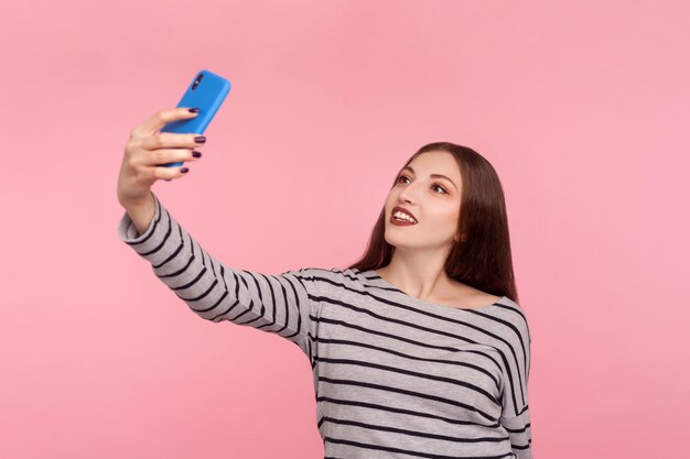 Retrato de una mujer hermosa con sudadera a rayas tomándose selfie en el teléfono móvil, sonriendo amablemente y hablando por videollamada, teniendo una conversación en línea. tiro de estudio interior aislado sobre fondo de color rosa