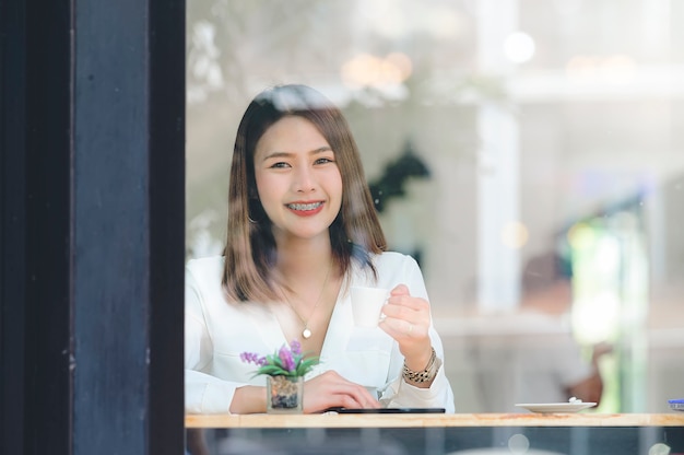 Retrato de mujer hermosa con taza de café, sonriendo y mirando a cámara mientras está sentado en el café.