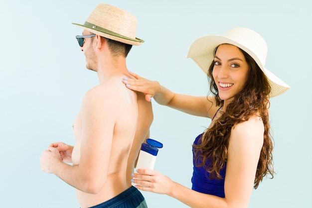 Retrato de una mujer hermosa en traje de baño sonriendo y haciendo contacto visual mientras aplica loción de protección solar en la espalda de un joven en la piscina