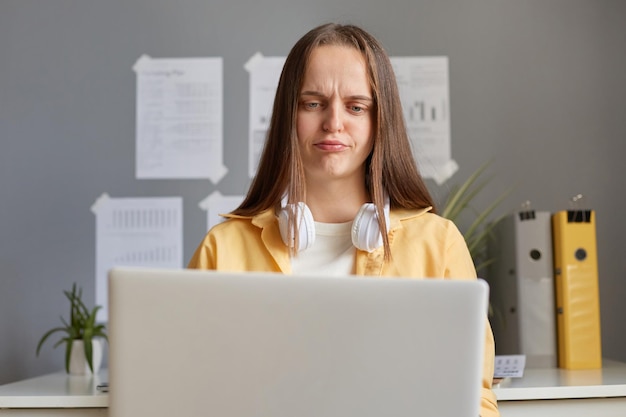 Retrato de una mujer hermosa y triste que usa una camisa amarilla sentada en la oficina usando su computadora portátil mirando la pantalla con el ceño fruncido expresando tristeza por no saber cómo hacer su trabajo