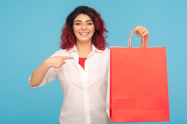 Retrato de mujer hipster adicta a las compras feliz con el pelo rojo de lujo apuntando grandes paquetes y sonriendo a la cámara disfrutando de las compras exitosas ventas de fin de semana tiro de estudio interior aislado sobre fondo azul