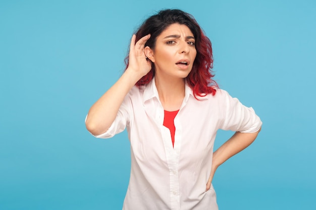 Retrato de mujer hipster entrometida con cabello rojo elegante en camisa escuchando atentamente sosteniendo la mano cerca de la oreja para escuchar una conversación confidencial, difícil de entender. Foto de estudio interior aislado sobre fondo azul.