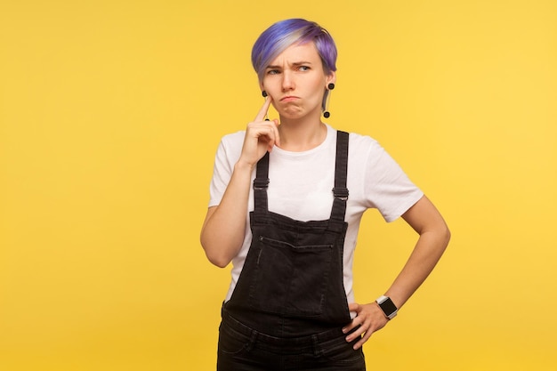 Retrato de una mujer hipster pensativa con el pelo corto teñido de violeta en overoles de mezclilla que se ve confundida, tiene dudas sobre cómo resolver el problema, piensa intensamente. aislado sobre fondo amarillo, foto de estudio