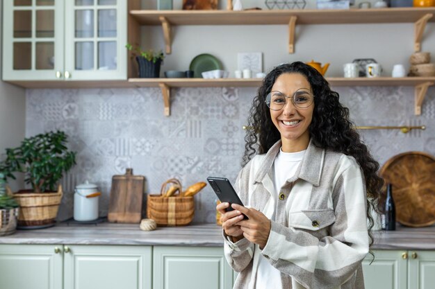 Retrato de mujer hispana en casa en la cocina mujer sosteniendo teléfono inteligente sonriendo y mirando a la cámara