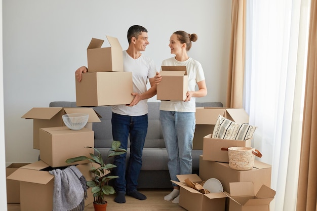 Retrato de una mujer y un hombre de familia feliz con pantalones blancos de pie con cajas de cartón que se mudan a una nueva casa desempacando pertenencias personales mirándose con amor inmobiliario