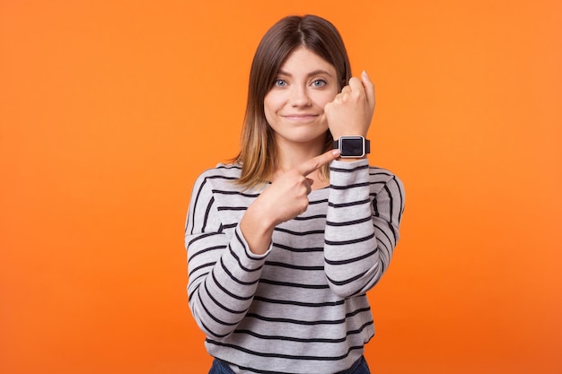 Retrato de una mujer impaciente positiva con cabello castaño en camisa a rayas de manga larga de pie señalando relojes en su muñeca mostrando el tiempo y sonriendo en un estudio interior aislado en un fondo naranja