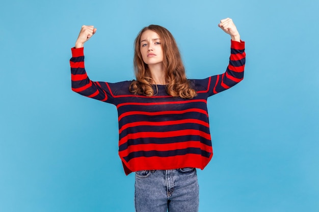 Retrato de mujer independiente confiada, mostrando bíceps, sintiendo energía, confianza y poder en las manos para ganar, tener éxito, usar suéter. Disparo de estudio interior aislado sobre fondo azul.