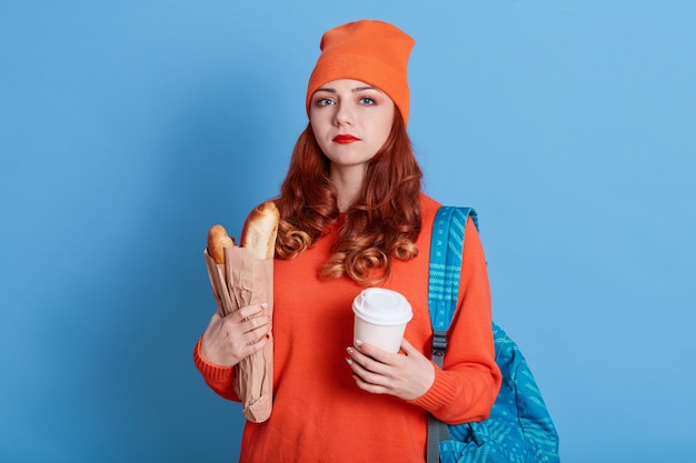 Retrato de mujer infeliz con sombrero y suéter casual, sosteniendo una bolsa de papel con baguettes y café para llevar