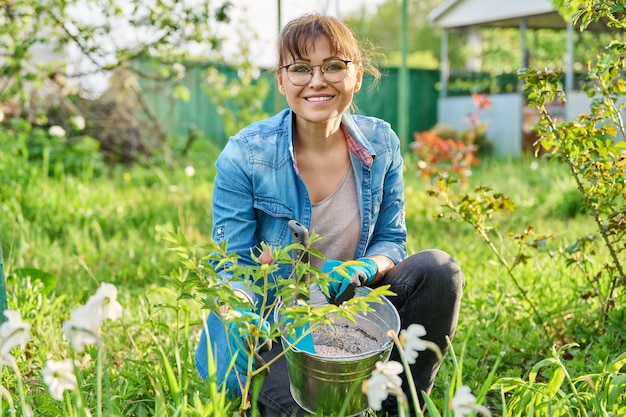 Foto retrato de una mujer en el jardín en el patio trasero fertilizando el lecho de flores de primavera con plantas con flores árbol peonía hermosa mujer mirando a la cámara sosteniendo un cubo de fertilizantes minerales jardinería paisajismo