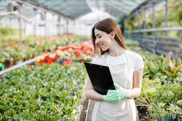 Retrato de mujer jardinera en el trabajo en invernadero con cuaderno examina las flores que crecen en invernadero Jardinería casera amor por las plantas y el cuidado Pequeña empresa