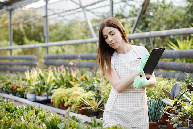 Retrato de mujer jardinera en el trabajo en invernadero con cuaderno examina las flores que crecen en invernadero Jardinería casera amor por las plantas y el cuidado Pequeña empresa