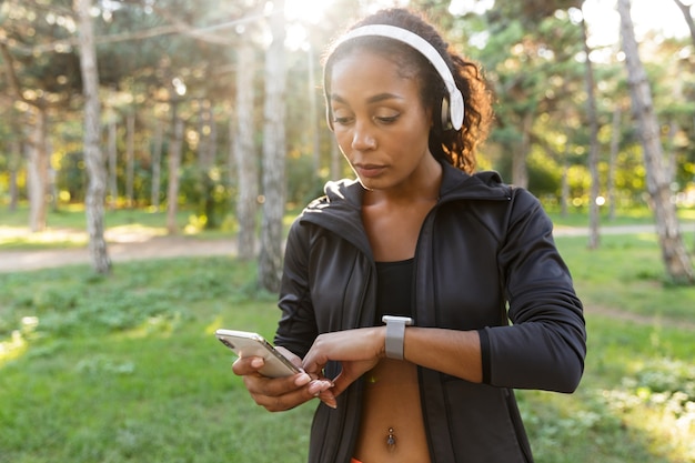 Retrato de mujer joven de 20 años vistiendo chándal negro y auriculares, mirando el reloj de pulsera mientras camina por el parque verde