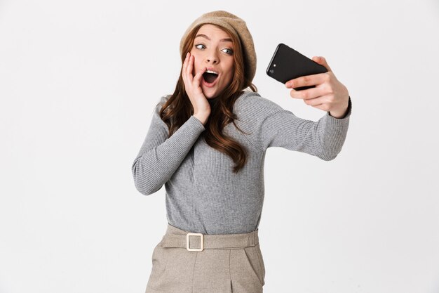 Retrato de mujer joven de 30 años con sombrero sonriendo y tomando selfie en teléfono celular aislado en blanco