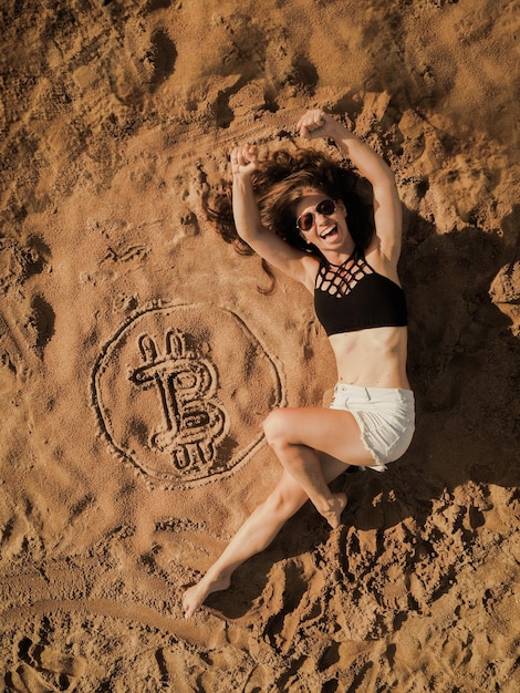 Foto retrato de una mujer joven acostada por el símbolo de bitcoin en la arena de la playa