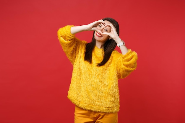 Retrato de mujer joven agradable en suéter de piel amarilla que muestra el corazón de forma con las manos aisladas sobre fondo de pared rojo brillante en estudio. Personas sinceras emociones, concepto de estilo de vida. Simulacros de espacio de copia.
