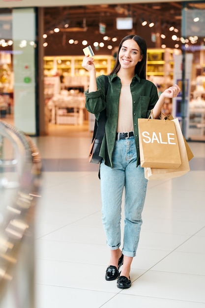 Retrato de mujer joven alegre y emocionada en traje casual con tarjeta de crédito y siendo recompensada por hacer compras en el centro comercial