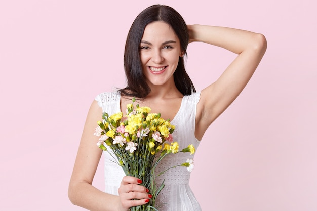 Retrato de una mujer joven alegre feliz en vestido blanco de verano con flores de colores en una mano, arreglando su cabello.