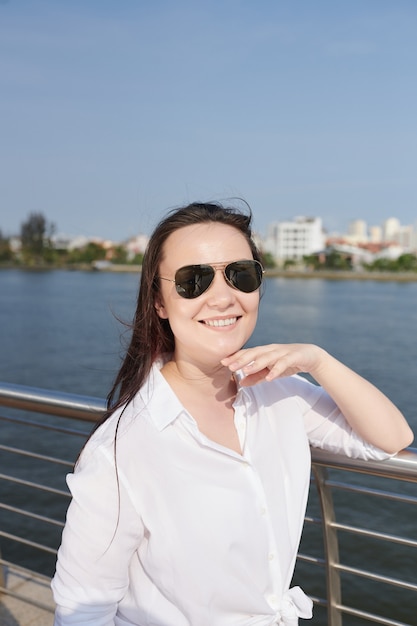 Retrato de mujer joven alegre en gafas de sol de pie en el puente y sonriendo a la cámara